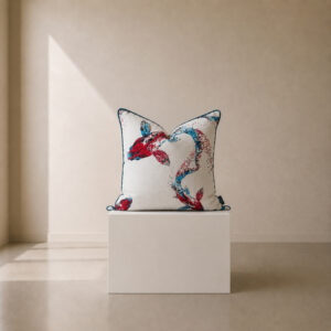Decorative scatter cushion featuring an artistic red and blue fish illustration on a soft ivory background, finished with contrasting teal piping and displayed on a minimalist white plinth.